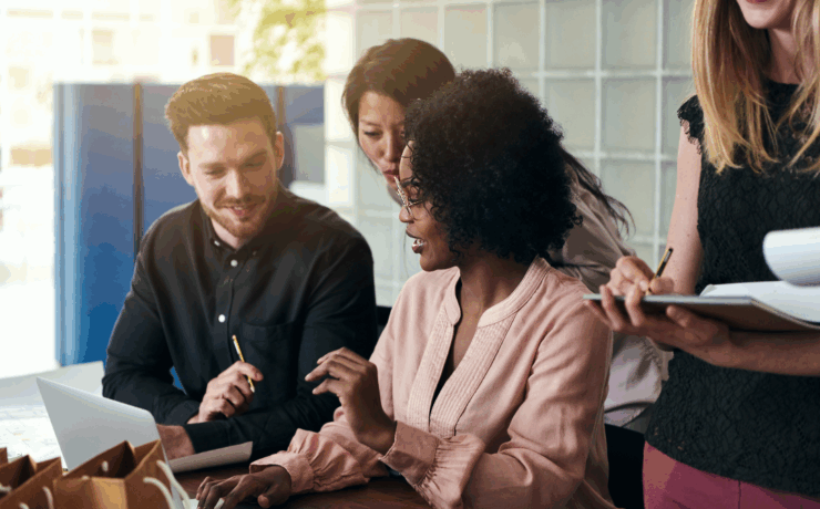 Group of diverse professionals collaborating around a laptop in a bright office setting, discussing ideas and taking notes.