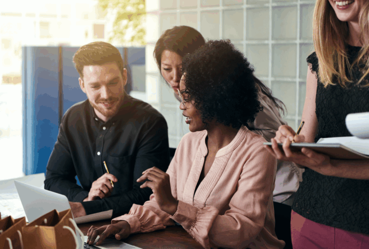Group of diverse professionals collaborating around a laptop in a bright office setting, discussing ideas and taking notes.