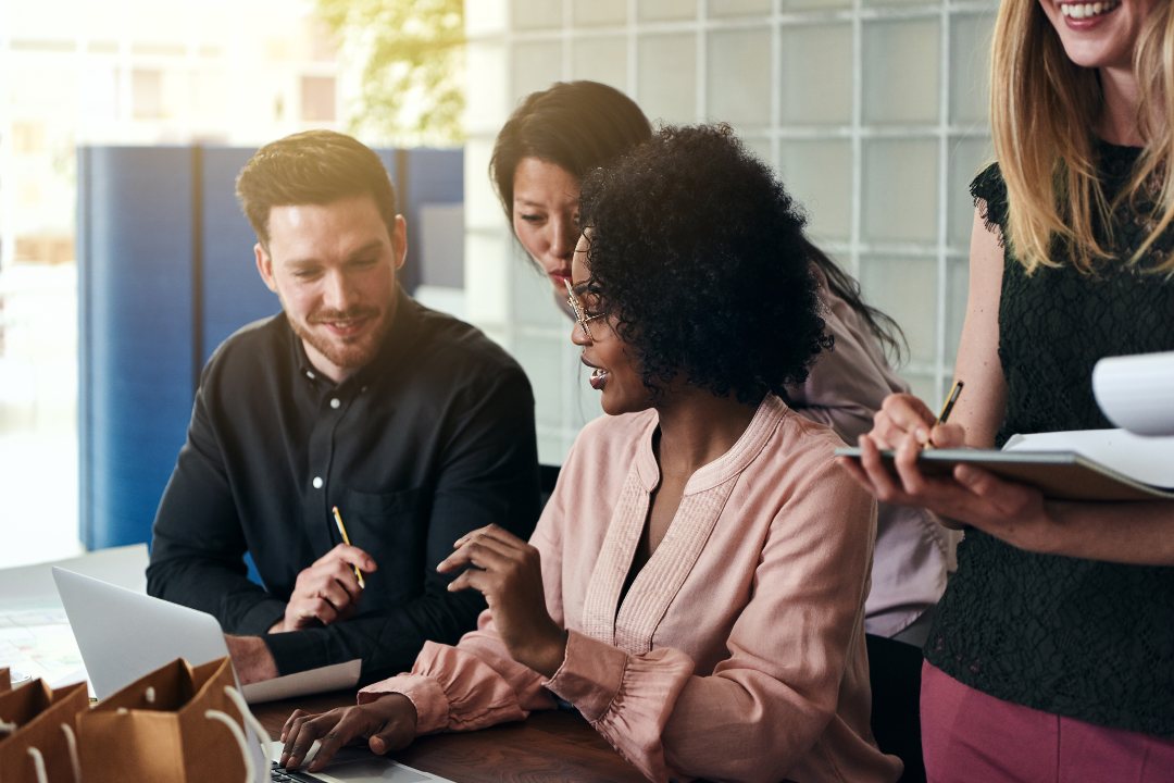 Group of diverse professionals collaborating around a laptop in a bright office setting, discussing ideas and taking notes.